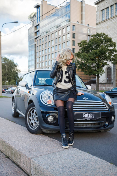 Outdoor portrait of beauty blond woman leaning on modern car. Stylish female model in trendy clothes standing close to little black car  - Фото, зображення
