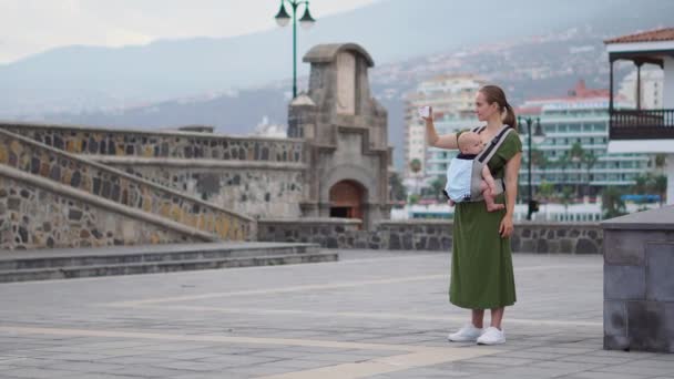Una giovane donna con un bambino in fionda viaggia in Europa. In piedi sulla piazza vicino all'oceano assume le attrazioni del telefono ed è in onda online per il tuo blog
 - Filmati, video