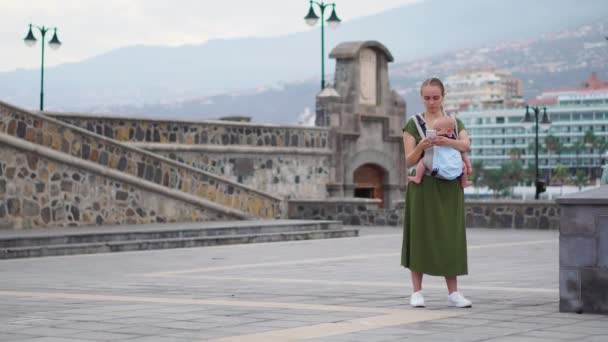 Nuori nainen, jolla on vauva kantositeessä, matkustaa Eurooppaan. Seisoo aukiolla lähellä merta vie puhelimen nähtävyyksiä ja on online-lähetys blogiisi
 - Materiaali, video
