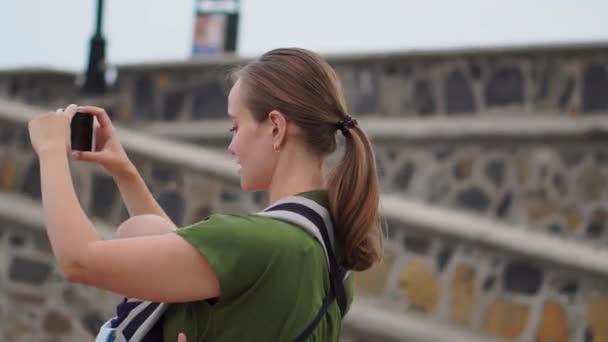 Una joven con un bebé en una honda viaja a Europa. De pie en la plaza cerca del océano toma las atracciones telefónicas y se transmite en línea para su blog
 - Imágenes, Vídeo