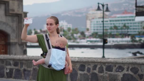 Μια νεαρή γυναίκα με ένα μωρό σε μια σφεντόνα ταξιδεύει στην Ευρώπη. Στέκεται στην πλατεία κοντά στον ωκεανό παίρνει για τα αξιοθέατα, τηλέφωνο και είναι μεταδίδονται σε απευθείας σύνδεση για το blog σας - Πλάνα, βίντεο