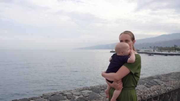 A young mom with a baby shoots on her hands looking at the ocean and the stunning view of the waves. Shows the child ships in the distant. - Πλάνα, βίντεο