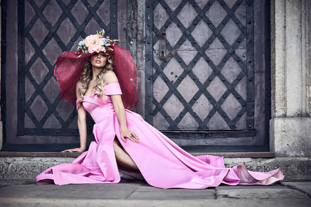 Full length portrait of pretty woman with light curly hair, in long pink silky dres, and big flower hat accessory with pink veil. Sitting near ancient door. Renaissance look. - Fotoğraf, Görsel