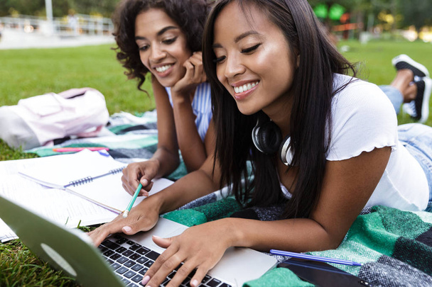 Photo of happy smiling young multiethnic friends girls doing homework using laptop outdoors in park. - Foto, immagini
