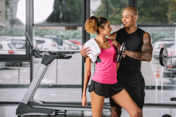tattooed african american sportsman embracing girlfriend with sport bottle and towel at gym - Foto, Imagem