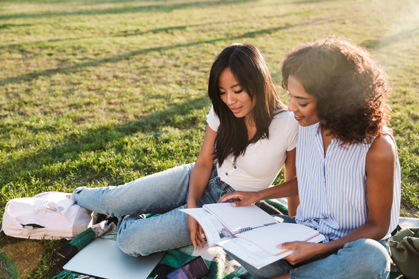 Duas jovens estudantes alegres sentadas em uma grama no campus, estudando com livros didáticos
 - Foto, Imagem