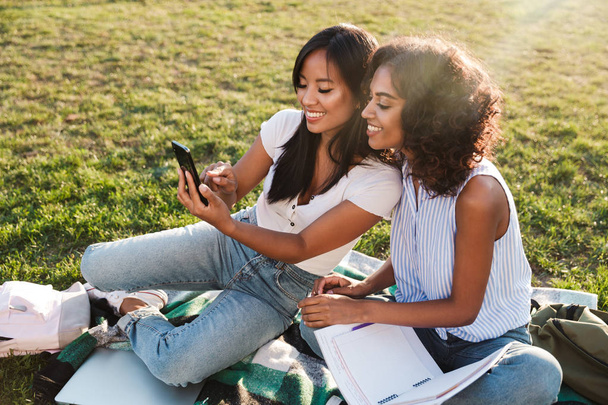 Two smiling young girls students sitting on a grass at the campus, studying with textbooks, taking a selfie - Foto, Imagem