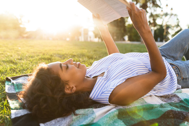 Улыбающаяся африканская девушка, лежащая на траве в парке, читающая книгу
 - Фото, изображение