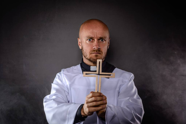 Καθολικός ιερέας στον Λευκό Λευκό ράσο και μαύρο πουκάμισο με γιακά κληρικός κρατώντας ξύλινο σταυρό και προσεύχεται - Φωτογραφία, εικόνα