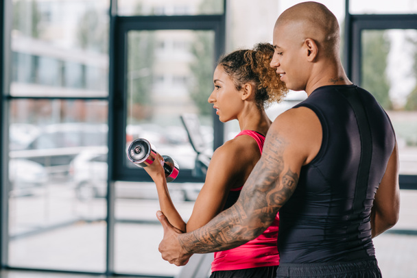 tattooed young african american male trainer helping sportswoman to exercising with dumbbell at gym - Photo, Image