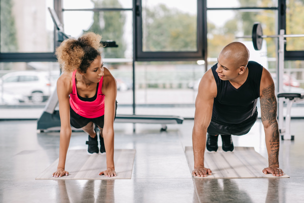 Afrikaanse Amerikaanse echtpaar van atleten kijken naar elkaar en plank op fitness matten bij sportschool doen glimlachen - Foto, afbeelding