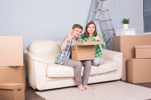 Недвижимость, переезд и переезд - молодая пара держит картонку, сидя на диване, и показывает вверх большие пальцы
 - Фото, изображение
