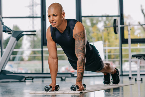 handsome african american sportsman with tattooed hand doing push ups with dumbbells on fitness mat at gym - Photo, Image