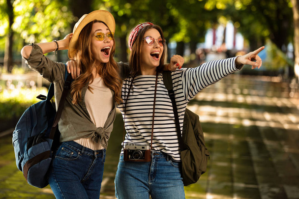 Image of excited pretty young happy women friends walking outdoors with backpacks hugging pointing. - Фото, изображение