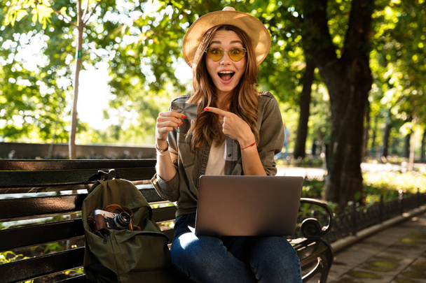 Photo of young beautiful excited happy woman outdoors sitting using laptop computer holding credit card pointing. - Photo, Image