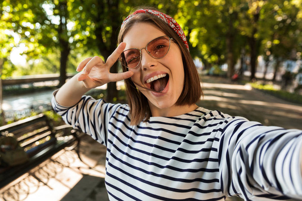 Photo of young beautiful excited happy woman outdoors take a selfie by camera showing peace gesture. - Zdjęcie, obraz