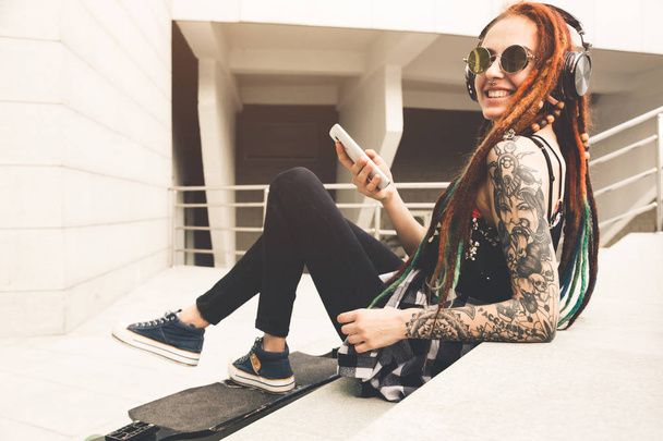 young girl with tattoo and dreadlocks listening to music while sitting on the steps - Foto, Imagen