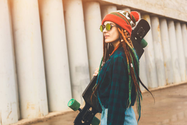 young girl in sun glasses and a cap stands with a longboard against the background of concrete structure - Fotografie, Obrázek