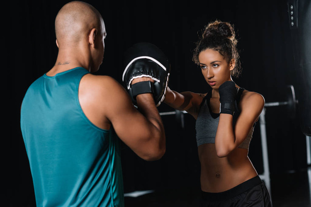 vista trasera del entrenador masculino muscular haciendo ejercicio con boxeadora femenina en el gimnasio
 - Foto, imagen