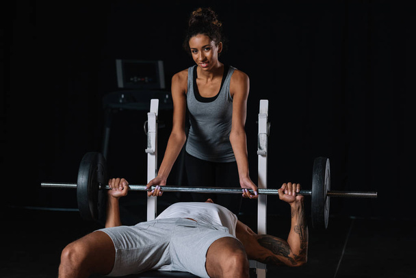 smiling african american woman helping boyfriend to exercising with barbell at gym - 写真・画像