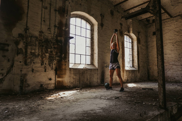 Atlética rubia haciendo kettlebell swing en un viejo edificio junto a la ventana
 - Foto, Imagen