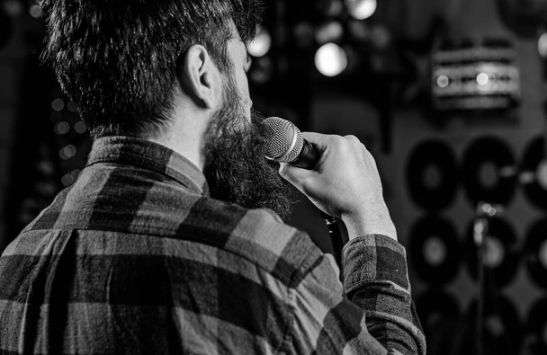 Musician with beard singing song in karaoke, rear view. Rock singer concept. Guy likes to sing in dark karaoke hall. Man in checkered shirt holds microphone, singing song, karaoke club background - Foto, Imagen