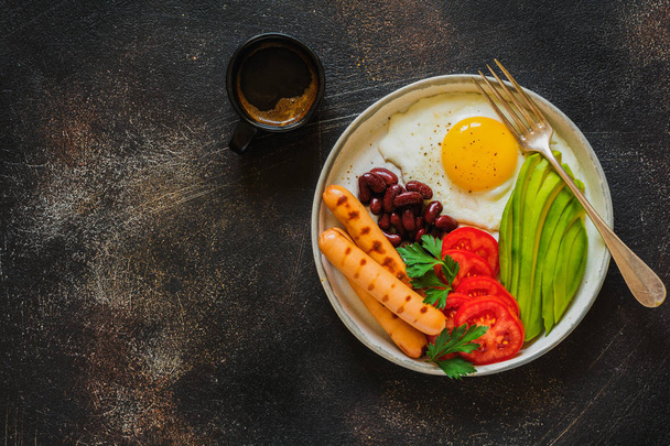 Traditional English breakfast with fried eggs, avocado, sausages grill, beans, and tomatoes on dark concrete background. Top view. - Photo, Image