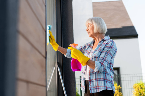 Elderly lady wearing bright yellow gloves feeling busy washing windows - Φωτογραφία, εικόνα