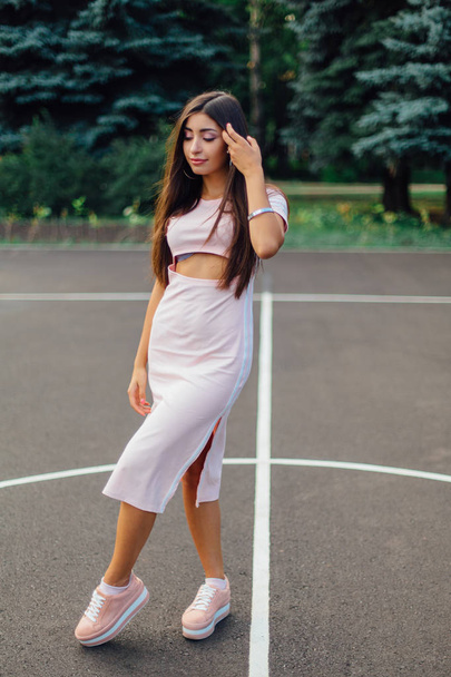 Charming brunette female dressed in a pink dress posing on the basketball court during sunset. - 写真・画像