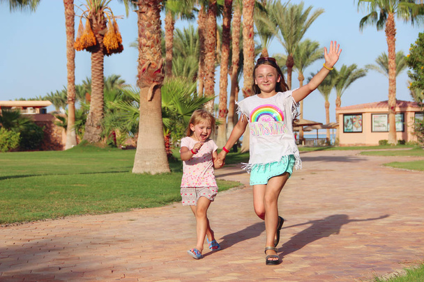 Счастливые сестры бегают по тропинке в тропическом курорте. Дети в Палм Парке. Радостные девушки бегут по тропинке среди финиковых деревьев. Дети наслаждаются праздниками. Сестры в отпуске. Девочки расслабляются
 - Фото, изображение