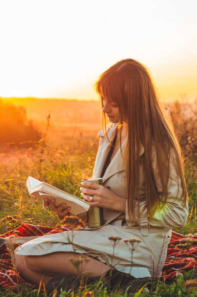 O conceito de estilo de vida recreação ao ar livre no outono. Menina com chapéu ler livros sobre xadrez com uma taça térmica. Outono. Pôr do sol. Aconchegante
 - Foto, Imagem