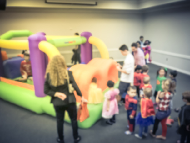 Motion blurred diverse multiethnic children enjoy inflatable bounce house jumping game. Defocused parents and kids at indoor school Halloween party in Texas, America - Photo, Image