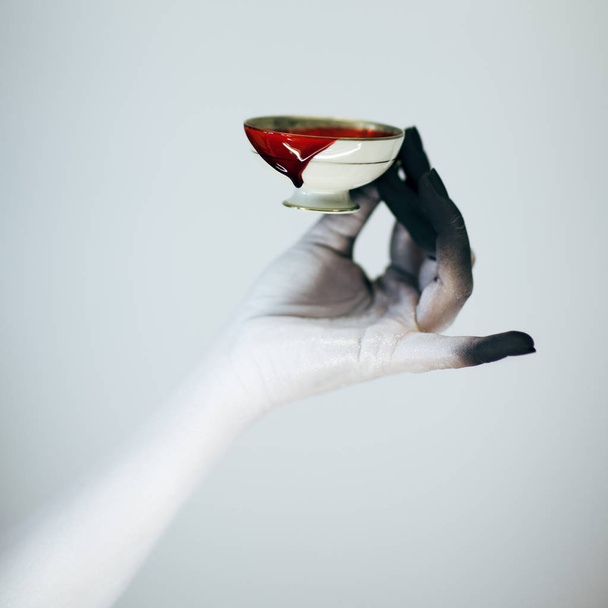 Creepy Halloween woman monster hand with white and black make up holding vintage cup with blood in front of white background - Photo, Image