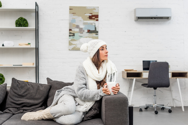 huomaavainen nuori nainen lämpimissä vaatteissa tilalla kuppi kuumaa teetä ja katselee pois, kun istuu sohvalla kotona
 - Valokuva, kuva