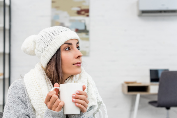 mujer joven reflexiva en ropa de abrigo sosteniendo taza de té caliente en casa y mirando hacia otro lado
 - Foto, Imagen