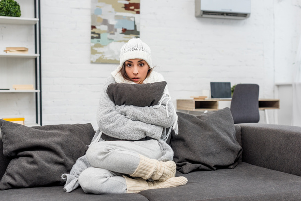 pakastettu nuori nainen lämpimissä vaatteissa istuu sohvalla ja halaus tyyny kotona
 - Valokuva, kuva