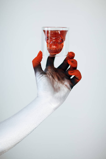 Creepy Halloween monster hand with white, red and black make up holding glass with skull of blood in front of white background - Foto, Imagem