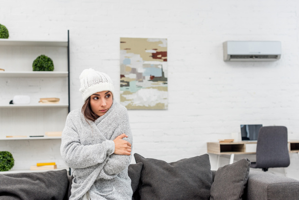 jäätynyt nuori nainen lämpimissä vaatteissa istuu sohvalla kotona ilmastointi roikkuu taustalla
 - Valokuva, kuva