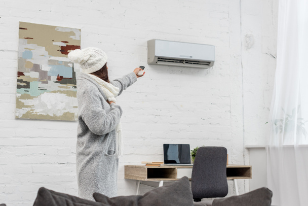 beautiful young woman in warm clothes pointing at air conditioner with remote control at home - Photo, Image