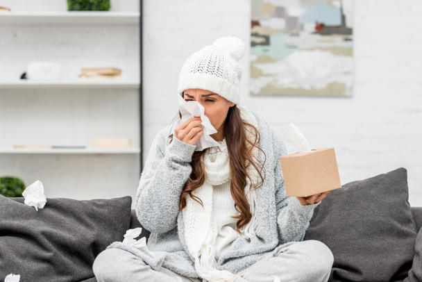 mujer joven enferma en ropa de abrigo sentada en un sofá desordenado y estornudando con servilletas de papel en casa
 - Foto, Imagen