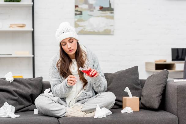 mujer joven enferma con ropa de abrigo vertiendo jarabe rojo para la tos mientras está sentada en el sofá en casa
 - Foto, Imagen