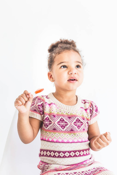 Portrait of a little girl in a colorful dress with a lollipop on a white background - Photo, Image