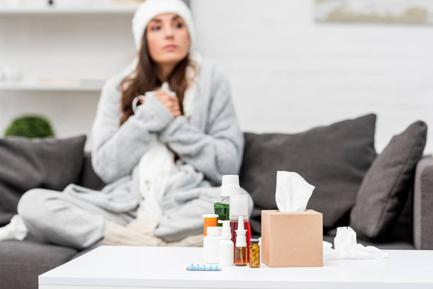 sick young woman in warm clothes sitting on couch at home with various medicines standing on table on foreground - Φωτογραφία, εικόνα