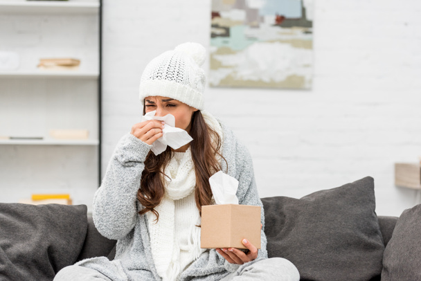sick young woman in warm clothes blowing nose with paper napkin at home - Photo, Image