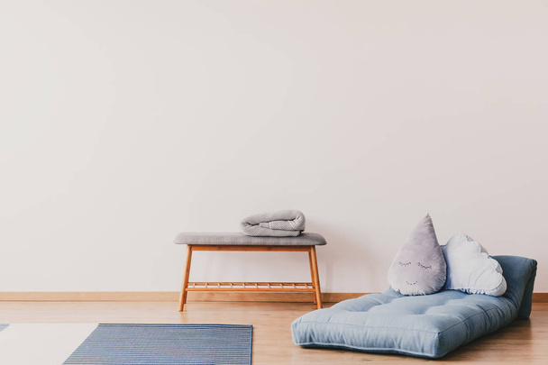 Blue futon with cute pillows on the floor of stylish baby room interior with copy space on the empty white wall and wooden and grey table with lamp - 写真・画像