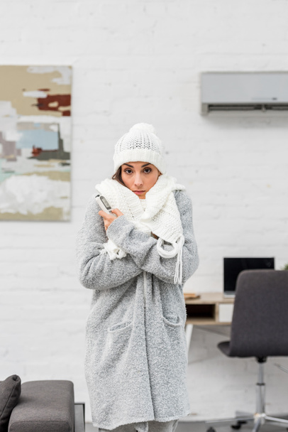 freezing young woman in warm clothes with air conditioner on background - Фото, изображение