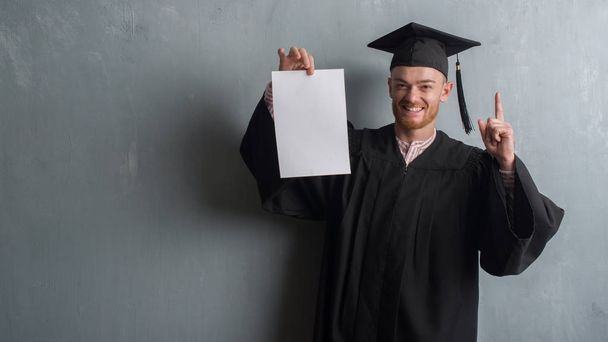 Young redhead man over grey grunge wall wearing graduate uniform holding degree surprised with an idea or question pointing finger with happy face, number one - Foto, imagen