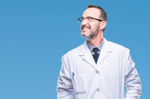 Middle age senior hoary professional man wearing white coat over isolated background looking away to side with smile on face, natural expression. Laughing confident. - Foto, imagen
