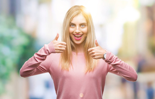 Young beautiful blonde woman wearing pink winter sweater over isolated background success sign doing positive gesture with hand, thumbs up smiling and happy. Looking at the camera with cheerful expression, winner gesture. - Photo, Image