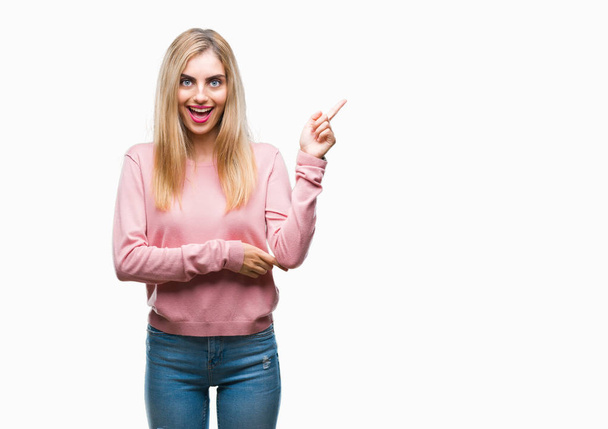 Jovem mulher loira bonita vestindo suéter de inverno rosa sobre fundo isolado com um grande sorriso no rosto, apontando com a mão e o dedo para o lado olhando para a câmera
. - Foto, Imagem